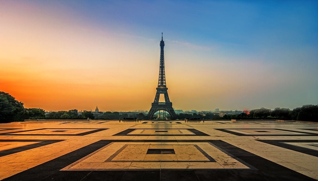 Torre Eiffel ao pôr do sol, vista de uma grande praça padronizada com um céu claro mudando de laranja para azul. Essa é a Place du Trocadéro, um dos lugares onde assistir o pôr do sol em Paris.