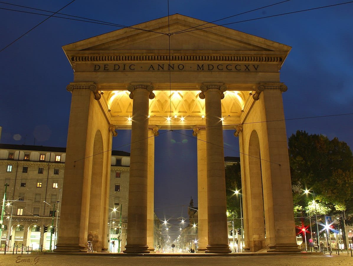 Porta Ticinese iluminada durante à noite. É um grande pórtico neoclássico com seis colunas contra um céu azul escuro. Prédios e postes de luz são visíveis.