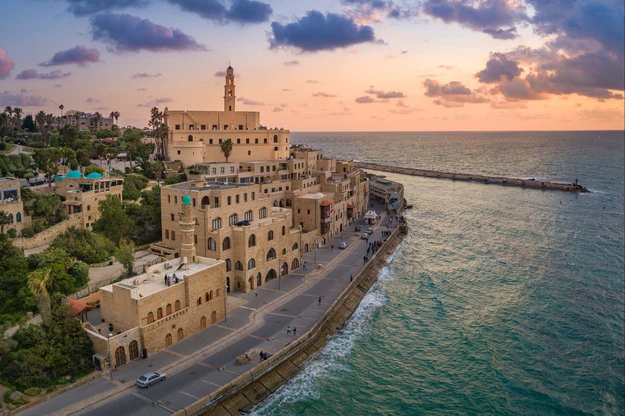 Vista de uma paisagem costeira do Porto de Jaffa (Tel Aviv) com edifícios históricos ao longo da orla ao pôr do sol, com vista para o mar e um céu nublado. Representa o post de o que fazer em Israel.