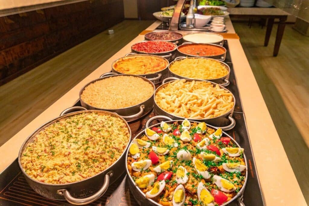 Uma variedade de pratos preparados em potes em uma mesa de bufê, incluindo massas, saladas e vários acompanhamentos, com um balcão de saladas ao fundo no Porto Seguro Praia Resort - All Inclusive.