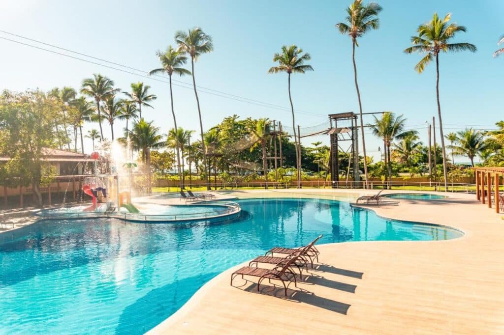 Um dia ensolarado no Porto Seguro Praia Resort - All Inclusive com uma grande piscina ao ar livre cercada por palmeiras e várias espreguiçadeiras no deck.