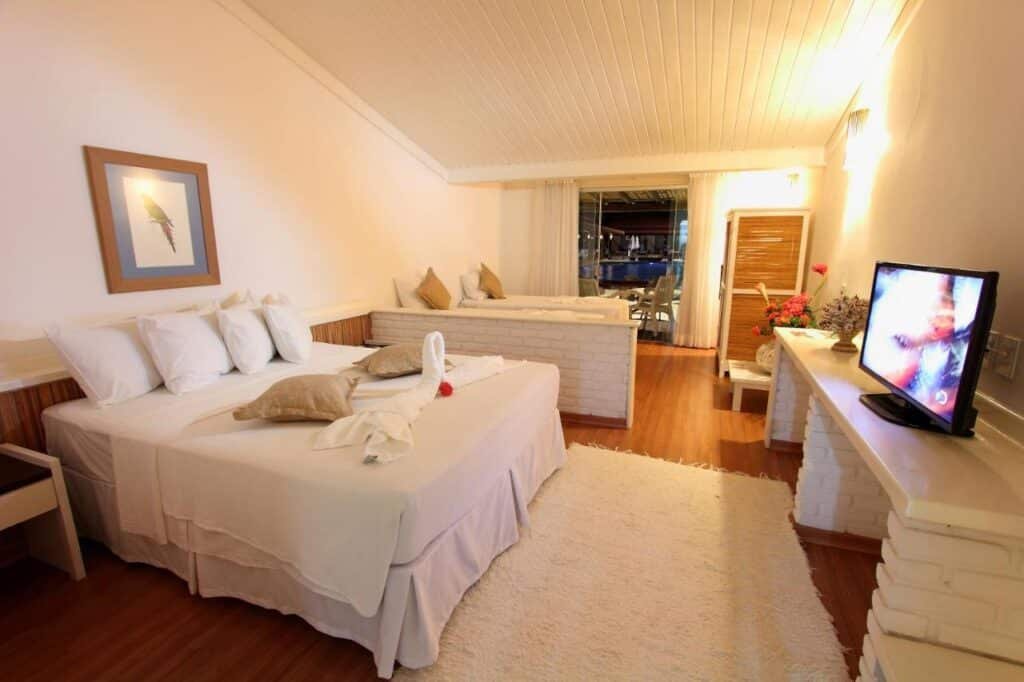 Quarto do Porto Seguro Praia Resort - All Inclusive com cama de casal, lençóis brancos, decoração de toalha de cisne, TV, móveis de madeira e portas de correr que levam a uma varanda.