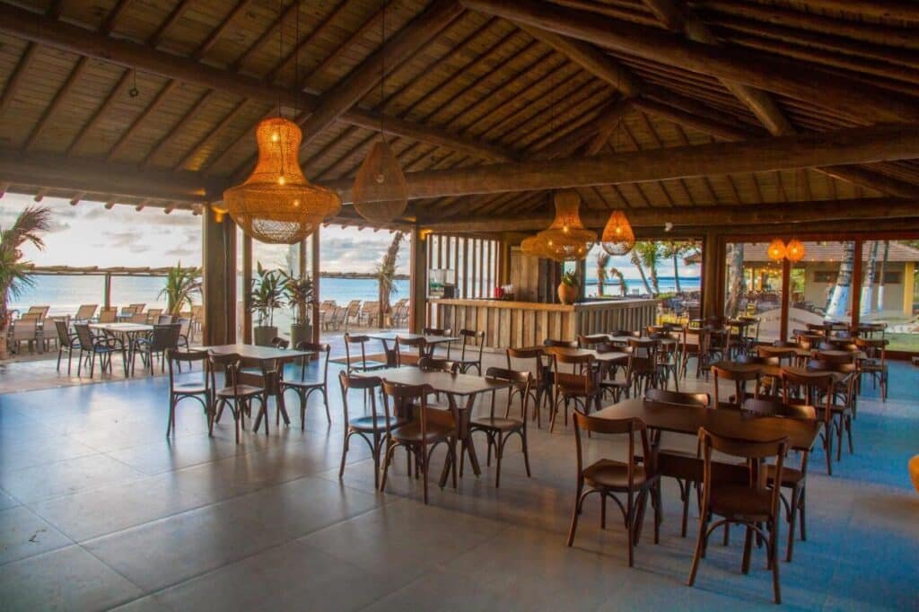 Restaurante ao ar livre do Porto Seguro Praia Resort - All Inclusive com mesas e cadeiras de madeira, luzes pendentes e vista para o oceano e palmeiras ao fundo.