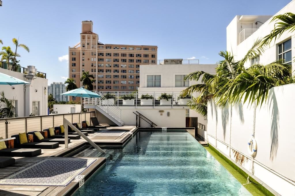 A imagem mostra uma piscina longa e estreita cercada por edifícios de estilo moderno no Posh South Beach, um dos hotéis em Miami Beach indicados. À esquerda da piscina, há uma área de descanso com espreguiçadeiras pretas e almofadas coloridas, parcialmente sombreada por guarda-sóis azuis. À direita, palmeiras decoram o espaço ao longo de uma parede branca. No fundo, é possível ver um edifício de vários andares em tons de bege. A atmosfera é ensolarada e relaxante, com um céu claro e azul acima, sugerindo um dia agradável.