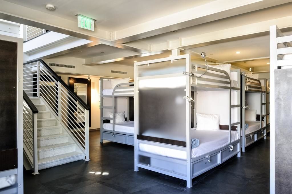 A imagem mostra um dormitório moderno com vários beliches metálicos, cada um equipado com um colchão branco, travesseiro e uma pequena luminária de leitura no Posh South Beach. À esquerda, uma escada de metal leva ao andar superior. O piso é escuro e a iluminação é suave, criando um ambiente limpo e organizado. As camas têm divisórias para privacidade e há uma sinalização de saída de emergência verde no teto. O espaço é funcional e bem planejado, ideal para acomodar várias pessoas confortavelmente.