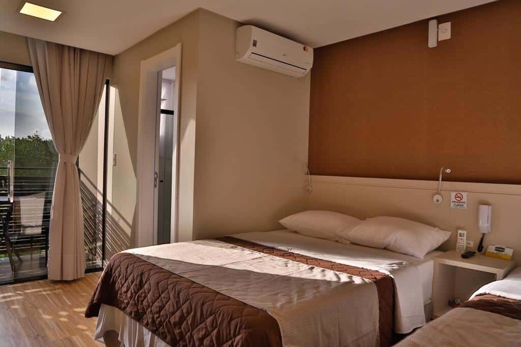 Quarto da Pousada Bella Casa, em Balneário Camboriú, com cama de casal, mesinha ao lado com telefone e controle do ar-condicionado, e uma varanda com mesa e vista para a natureza verde