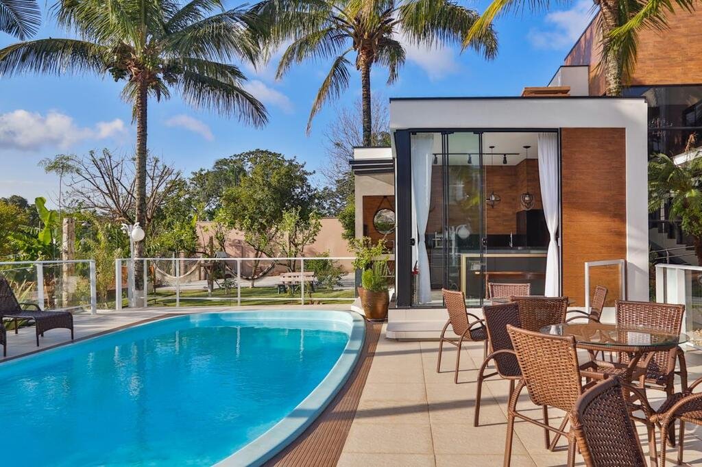 Área externa com piscina da Pousada Bella Casa, sendo do lado esquerdo uma piscina azul oval com algumas espreguiçadeiras em volta, e no lado direito há algumas mesas redondas com cadeiras e uma área gourmet de cozinha. Ao fundo há um jardim com redes e árvores