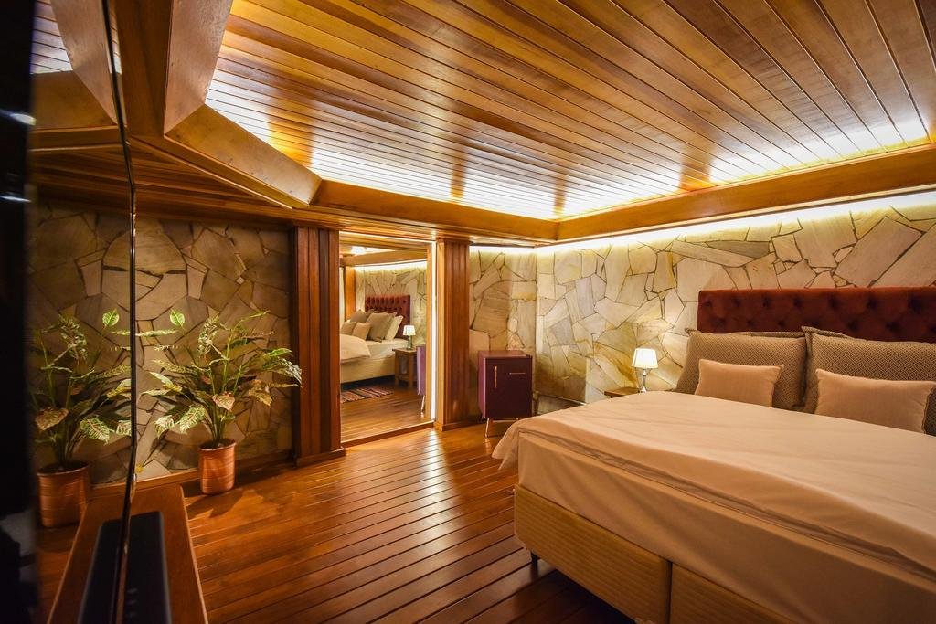 suíte da Pousada Borô em Gramado com uma cama de casal à direita e uma televisão de tela plana à esquerda. O quarto conta com paredes de pedra desenhadas e teto e chão de madeiras.