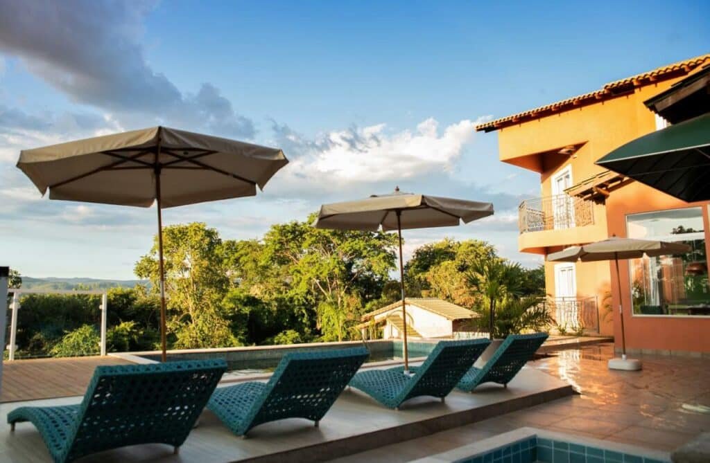 Área externa piscina com cadeiras de praia e guarda sol, fachada do hotel ao lado com as sacadas dos quartos. Piscina de borda infinita com vista para vegetação em um dia de céu azul com algumas nuvens. 