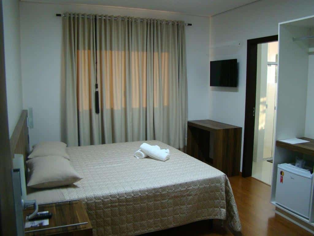 Interior do quarto da Pousada by Rieger com uma cama de casal, frigobar, Tv e um mesa pequena de maadeira