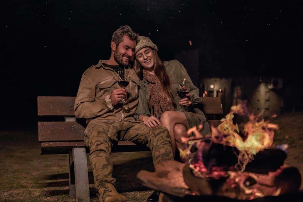 Um casal está sentado em um banco, sorrindo e segurando taças de vinho, em frente a uma fogueira à noite, sob um céu estrelado para representar um dos melhores lugares para lua de mel