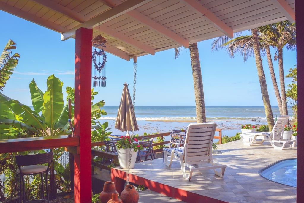 Varanda à beira-mar da Pousada Canto da Brava, com uma piscina, espreguiçadeiras e mesas com cadeiras em volta e bem a frentem tem o mar com ondas pequenas, em um dia ensolarado e com poucas nuvens