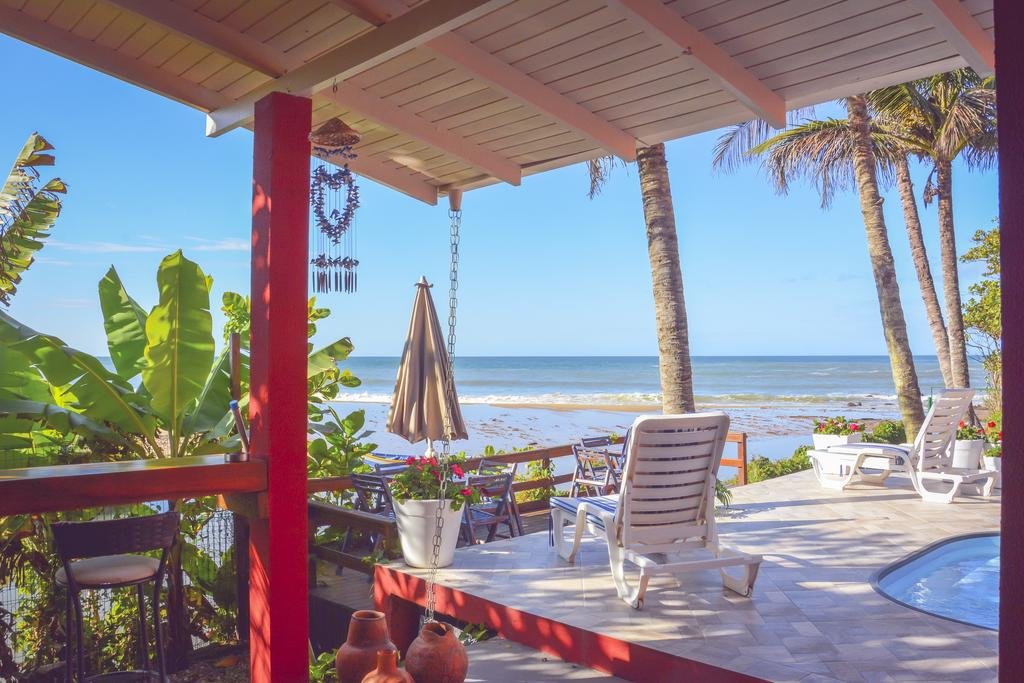 Varanda à beira-mar da Pousada Canto da Brava, em Balneário Camboriú, com uma piscina, espreguiçadeiras e mesas com cadeiras em volta e bem a frentem tem o mar com ondas pequenas, em um dia ensolarado e com poucas nuvens