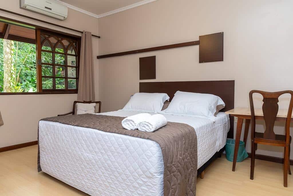 Quarto da Pousada Casa da Pedra, com uma cama de casal com dois travesseiros e duas toalhas enroladas brancas em cima, uma mesa com uma cadeira de madeira ao lado, uma poltrona do outro lado e uma janela com vista da natureza, composta por folhas verdes, no lado de fora