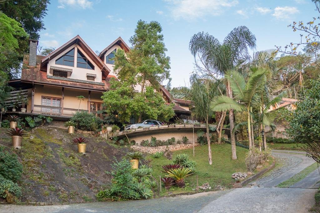 Frente da Pousada Casa da Pedra, com um caminho dando acesso ao sobrado, que fica em uma parte elevada e em cima de uma pedra, que tem jardim, vasos de flores e árvores ao lado