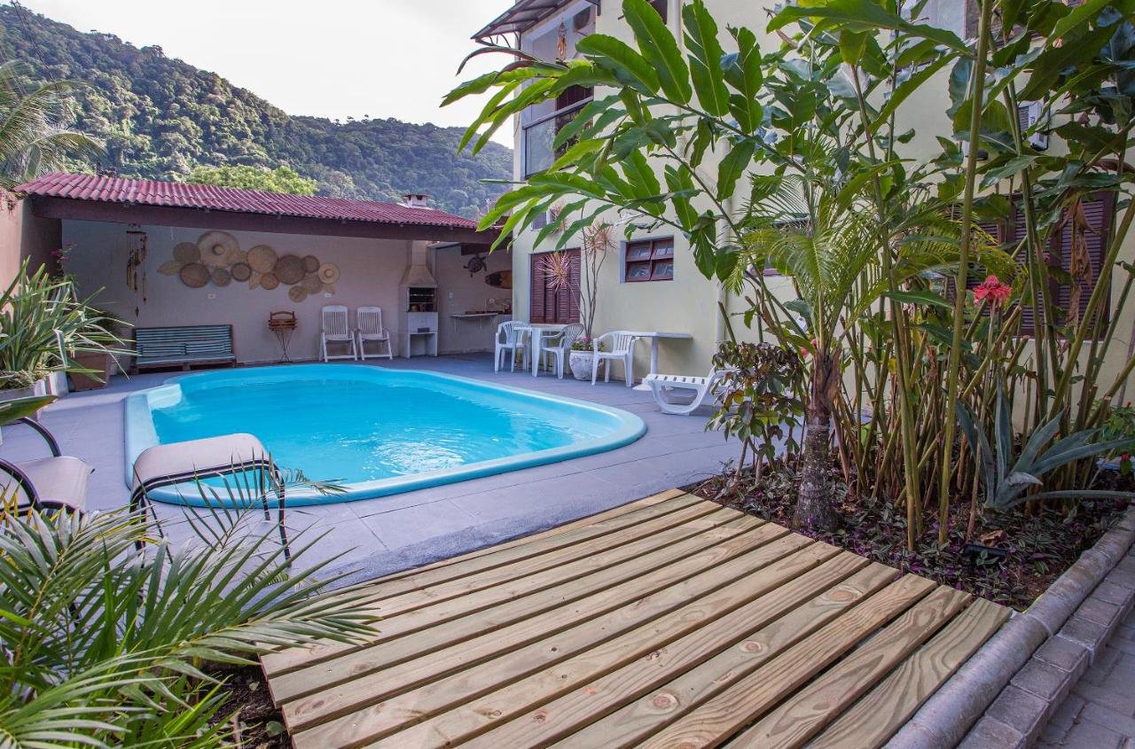 Área de piscina ao ar livre com espreguiçadeiras na Pousada Castelinho, cercada por plantas tropicais e um deck de madeira, ao lado de uma casa com assentos no pátio.