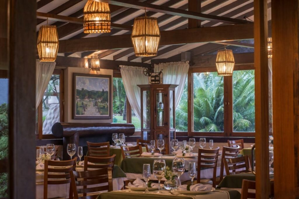 Área do restaurante na Pousada Cavaleiro dos Pirineus, com mesas já postas com guardanapo de pano, pratos, taças e janelas ao entorno com vista para vegetação. 