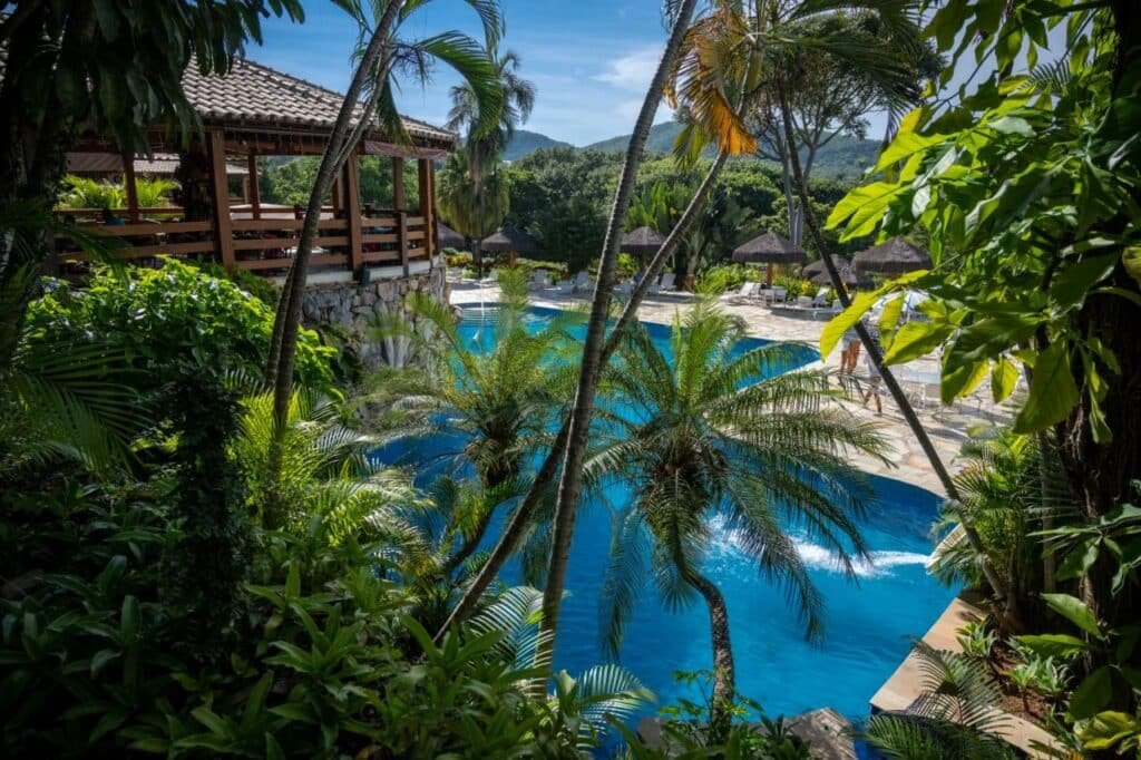 Resort em Pirenópolis, área externa da piscina cercado de vasta vegetação. Ao fundo cadeiras de praia e guarda sol. 