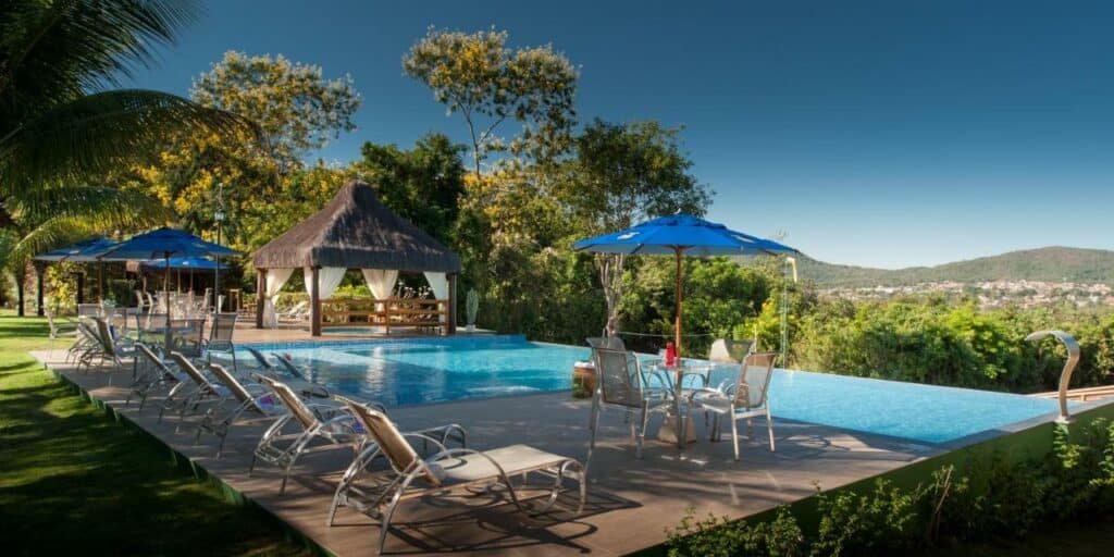 Área da piscina na Pousada Cavaleiro dos Pirineus com vista para a cidade, piscina de borda infinita com cadeiras e guarda-sol em volta. 