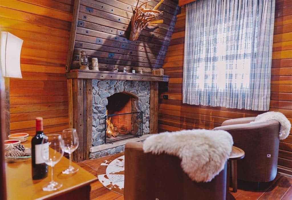 Sala na Pousada Chalés Por do Sol com duas poltronas com cobertas felpudas, uma lareira, um mesinha com vinho e taças, além de uma janela com cortinas