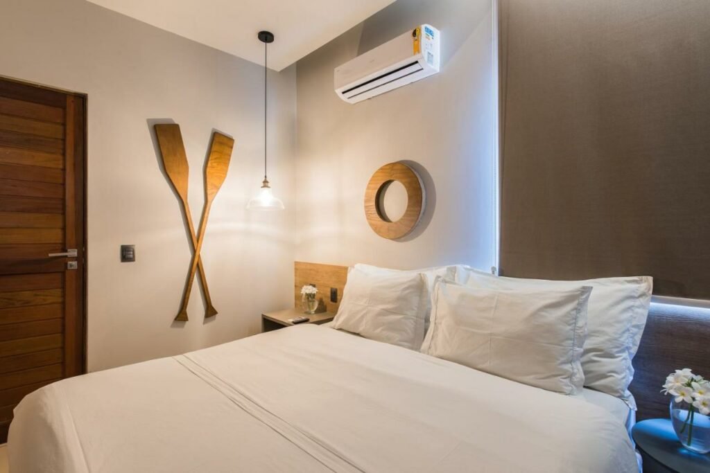 Quarto de casal na Pousada Corveta, com cama ampla, quatro travesseiros, fronhas e lençol brancos, enfeite de remos na parece e ar-condicionado e cortina blecaute sobre a área de cabeceira
