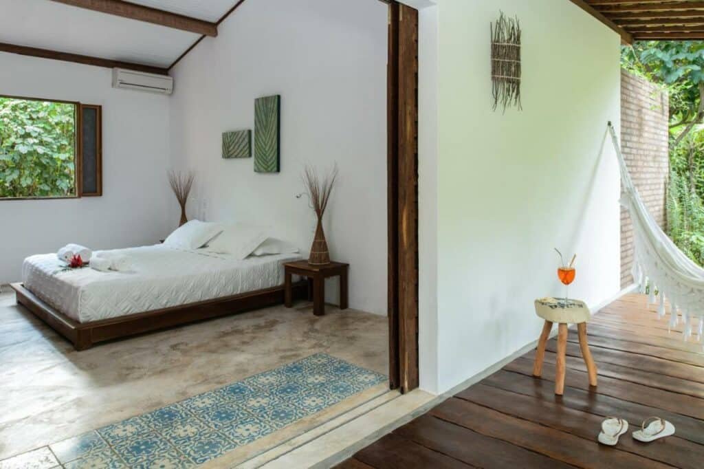 Quarto de casal na Pousada da Amendoeira, com cama baixa, amplo espaço, mesinhas de cabeceira com enfeite, e uma área externa com rede de descanso