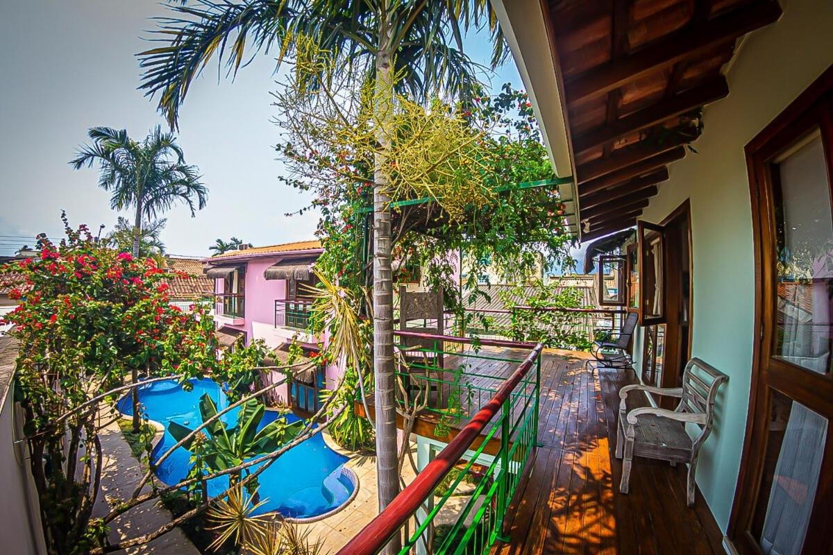 Vista da varanda de um quintal tropical com piscina, cercado por plantas e árvores exuberantes, e uma cadeira no chão de madeira da varanda. Essa é a Pousada da Banda, uma das recomendações de pousadas em São Sebastião.