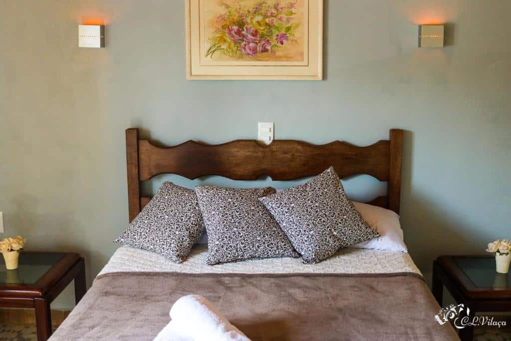A imagem mostra uma cama de casal com uma cabeceira de madeira escura em um quarto aconchegante. Sobre a cama, há três travesseiros decorativos com estampas florais em tons de preto e branco, além de uma manta marrom cobrindo os lençóis brancos. Na parede atrás da cama, há um quadro com uma pintura de flores em cores suaves, complementando a decoração. Ao lado da cama, dois criados-mudos com tampo de vidro seguram pequenos objetos decorativos. A iluminação suave nas paredes cria uma atmosfera tranquila e convidativa no ambiente. Essa é a Pousada e Restaurante Victórios, um dos hotéis-fazenda em Minas Gerais.