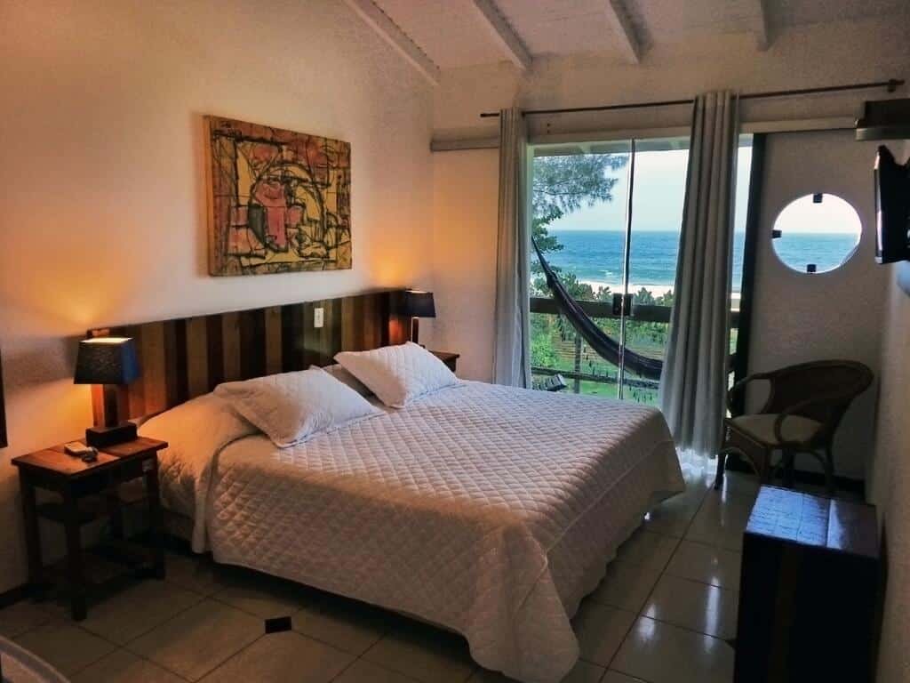 Quarto da Pousada Estaleiro Village, com cama e casal, uma mesinha de cada lado com um abajur em cima, uma cadeira ao lado direito e uma TV suspensa na parede. Há também uma porta de vidro dando acesso à sacada, que tem uma rede e vista para a natureza e praia, para ilustrar as pousadas em Balneário Camboriú
