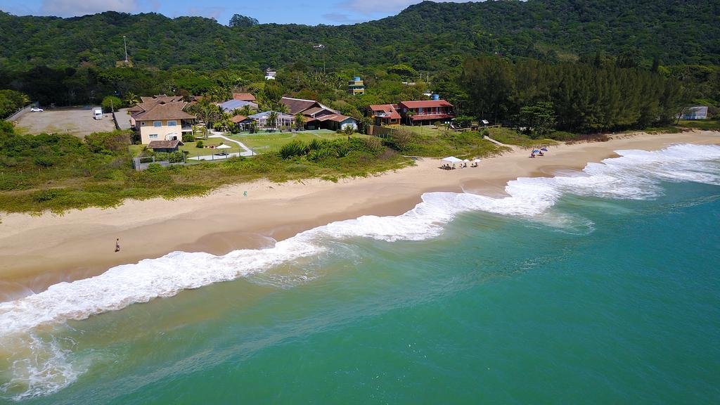 Praia com água esverdeada cristalina e areia bege, com algumas pessoas andando e outras sentadas em baixo de guarda-sóis, com casas em uma área cheia de natureza, sendo uma delas a Pousada Estaleiro Village