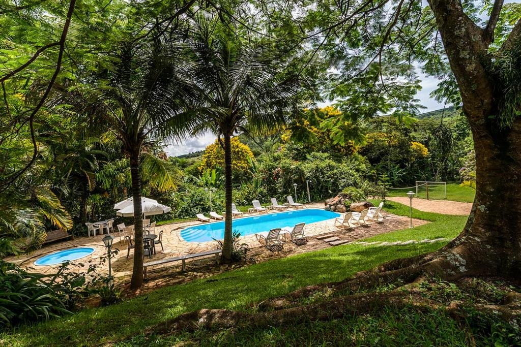 Uma pequena piscina ao ar livre cercada por espreguiçadeiras e mesas com guarda-sóis, situada em um jardim exuberante com árvores e vegetação no Pousada Famiglia Bartho, um dos hotéis fazenda em São Paulo.