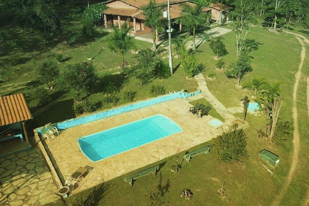 A imagem mostra uma vista aérea de uma propriedade rural com uma piscina retangular cercada por um piso de pedra. Ao redor da piscina, há algumas espreguiçadeiras e um espaço para relaxar. A área ao redor é verde, com gramado e várias árvores espalhadas pelo terreno. Ao fundo, há uma casa simples com telhado de cerâmica e varanda. Um caminho de pedra leva da piscina até a casa, e o ambiente parece tranquilo, em meio à natureza, ideal para momentos de lazer ao ar livre. Essa é a Pousada Fazenda Bocaina, um dos hotéis-fazenda em Minas Gerais.