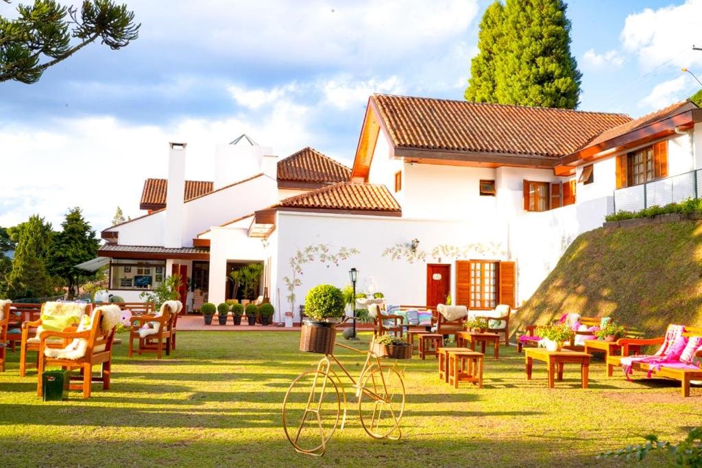 Jardim da Pousada Hortelã com mesas e cadeiras de madeira, alguns vasos de plantas e a propriedade é branca com janelas de madeira