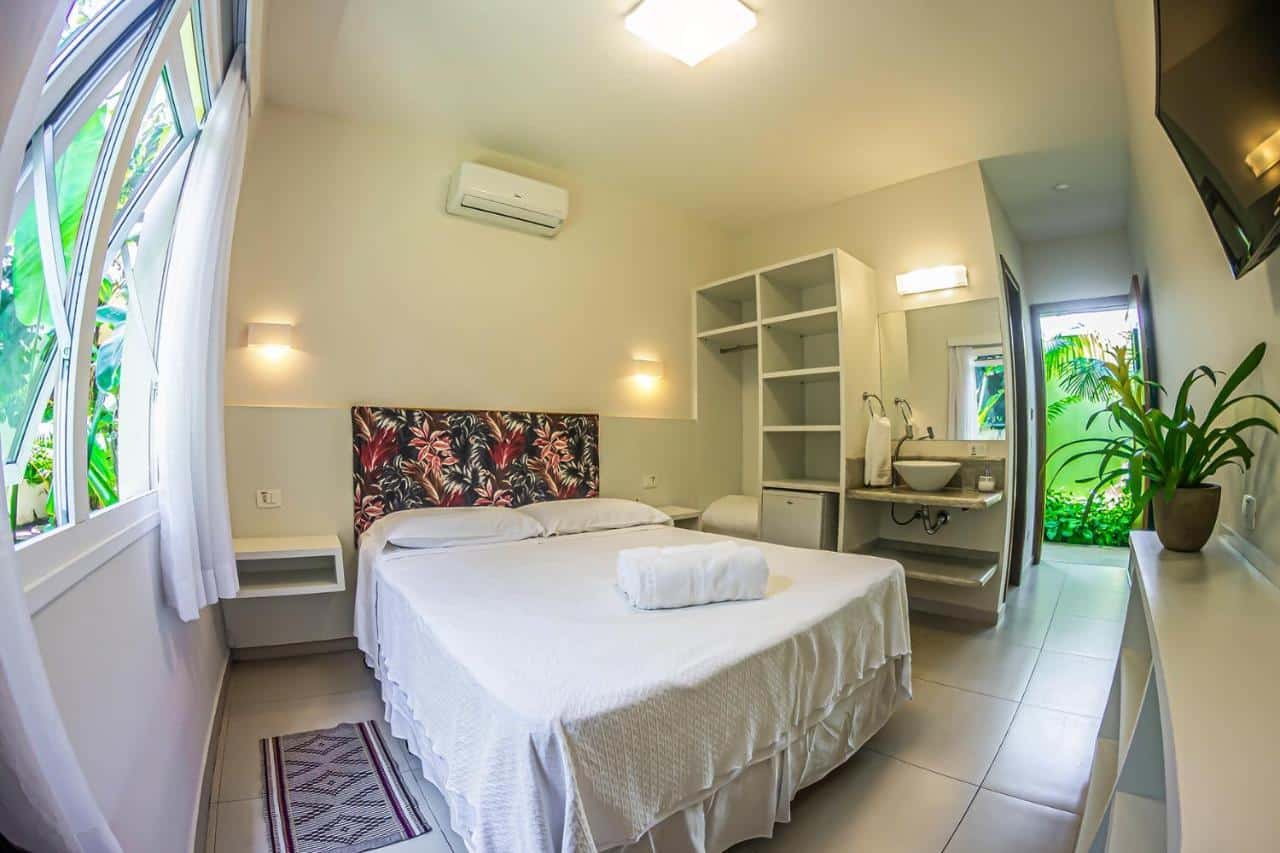 Um quarto pequeno e moderno com uma cama de casal, roupa de cama branca, ar condicionado e uma pia de banheiro visível através de uma porta aberta. Essa é a Pousada Ipê, uma das recomendações de pousadas em São Sebastião.