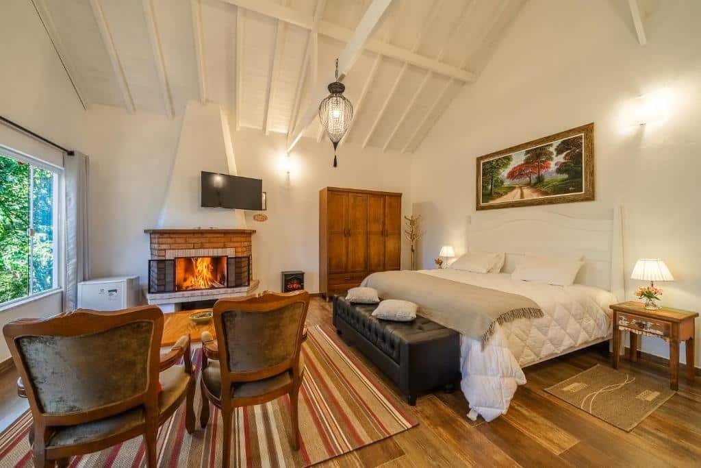Quarto aconchegante da Pousada Jardim da Mantiqueira com lareira, cama, poltronas de madeira e TV. Um tapete colorido cobre o piso de madeira. Arte emoldurada na parede.