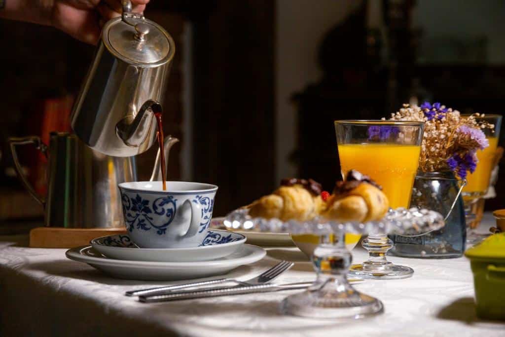 A foto mostra uma cena de café da manhã. Uma pessoa está servindo café de uma cafeteira prateada em uma xícara azul e branca com padrão. A mesa está posta com vários itens de café da manhã, incluindo croissants, um copo de suco de laranja, talheres e pratos. Há também elementos decorativos, como flores, que adicionam um toque especial ao ambiente. A imagem captura a sensação de conforto e aconchego de uma rotina matinal, destacando o prazer simples de um café da manhã bem preparado.