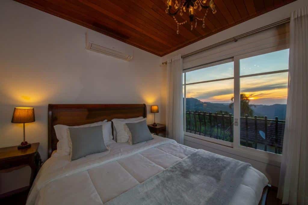Foto do interior do Pousada La Vista representando o post de chalés em Gramado. O quarto deste chalé é bastante acolhedor e elegante. Ele possui uma cama grande com uma cabeceira de madeira, arrumada com roupa de cama branca e almofadas cinza. Ao lado da cama, há mesas de cabeceira com abajures que emitem uma luz quente. Acima da cama, há um ar-condicionado e o teto é revestido com painéis de madeira, o que adiciona um toque rústico ao ambiente. Um lustre grandioso pende do centro do teto.

O quarto também conta com uma grande janela na didreita que oferece uma vista deslumbrante das montanhas e árvores ao entardecer, sugerindo que o chalé está localizado em uma área cênica.