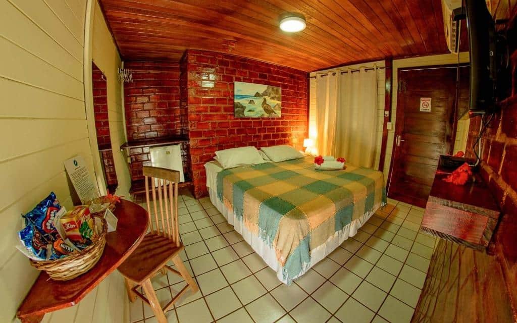 Quarto de casal na Pousada Lenda das Águas, com cama box, frigobar, mesa com cesta cheia de doces e salgadinhos, TV na parede, e cortina na janela