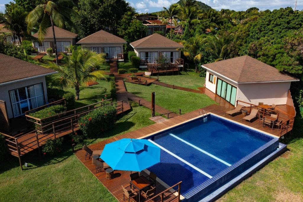 Chalés em volta da piscina da Pousada Lua Bela, em Fernando de Noronha, onde há um deck com espreguiçadeiras e um guarda-sol, além de muito verde e gramado pelo terreno