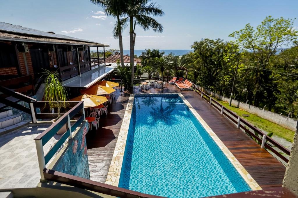Área externa com piscina da Pousada Magnus, com uma piscina retangular, mesas com guarda-sóis e espreguiçadeiras ao redor. No lado direito há árvores e no fundo uma vista para o mar azul