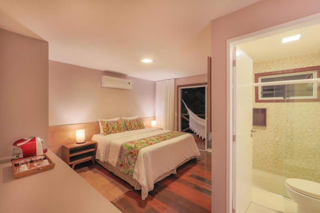Quarto de casal na Pousada Mar Atlântico, com cafeteira no canto esquerdo, cama com mesa de cabeceira e abajures, ar-condicionado, banheiro com box de vidro, e cortina na frente da varanda com rede de descanso