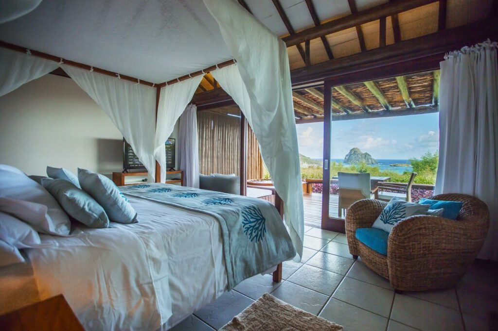 Quarto de casal com dossel sobre a cama, poltrona com almofadas no canto direito, mesa com TV plana na parede do lado esquerdo, além de varanda com vista para o oceano