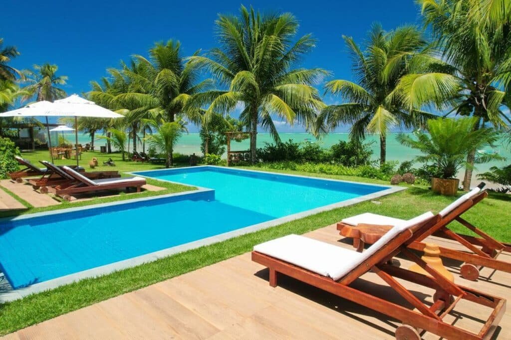 Piscina da Pousada Marceneiro, com espreguiçadeiras posicionadas em volta, gramado e alguns coqueiros, além do mar turquesa de Milagres bem pertinho