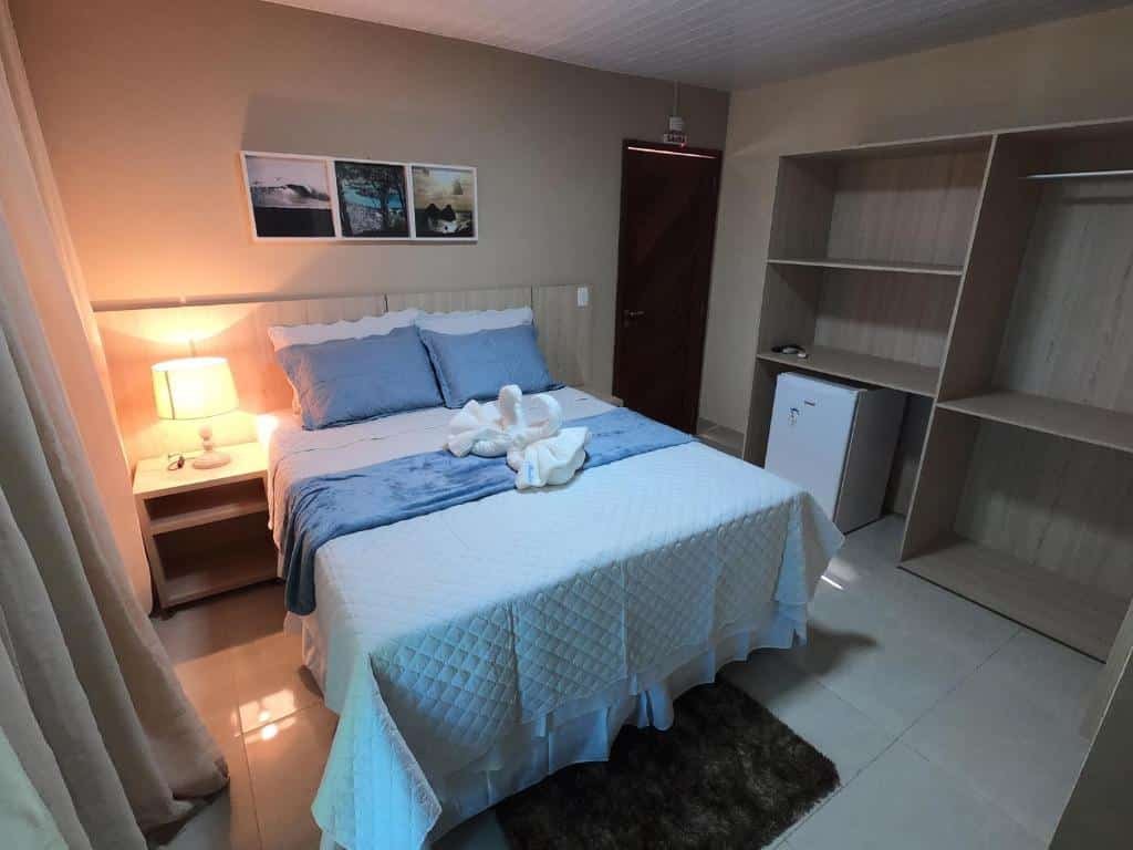 Quarto de casal da Pousada Marlin Azul, com frigobar embaixo do armário, três quatros na parede sobre a cama, e um abajur aceso na mesinha de cabeceira