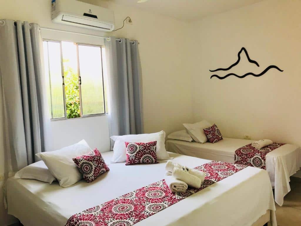 Quarto com uma cama de casal e uma cama de solteiro na Pousada Morro do Pico. Há uma janela sobre a cama, com cortina, e a logo da pousada na parede sobre a outra cama