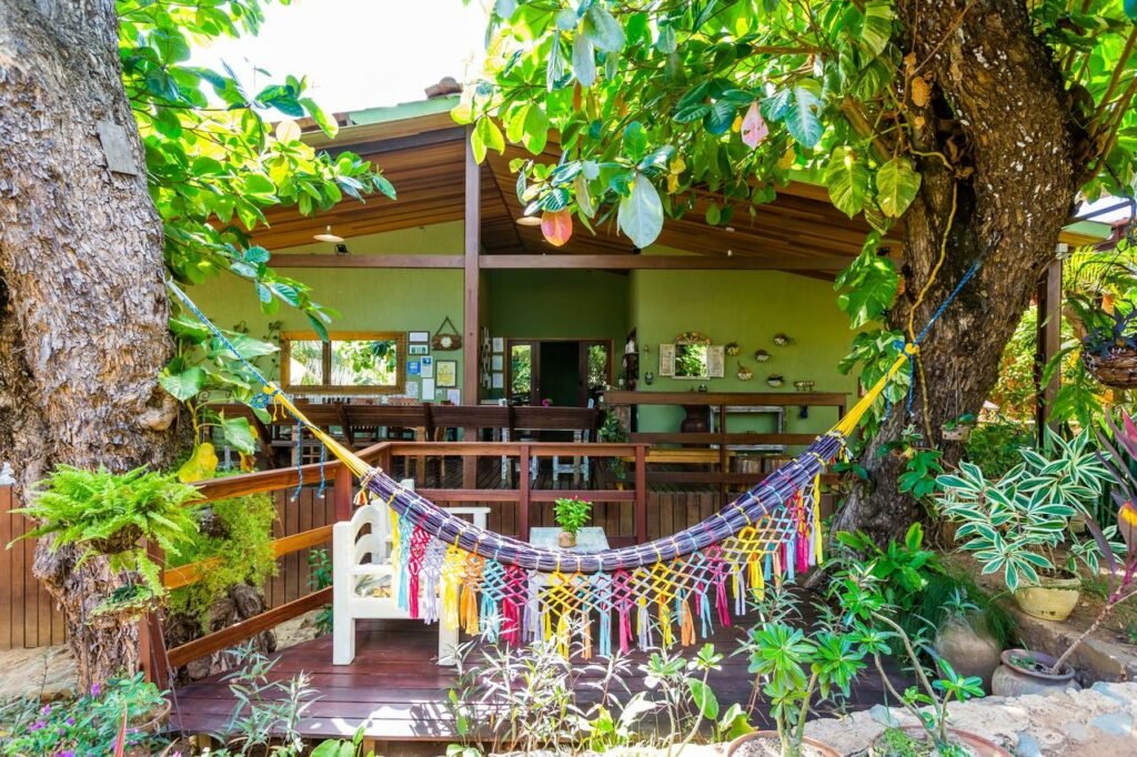 Rede de descanso colorida pendurada em árvores do jardim da Pousada Naiepe, em Fernando de Noronha, com parte da casa de hospedagem ao fundo, de paredes verdes