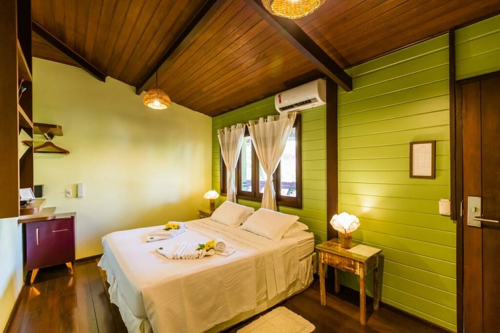 Quarto da Pousada Naiepe, com cama de casal, janela na parede sobre a cama, ar-condicionado, mesinhas de cabeceira com abajures, e um frigobar no canto