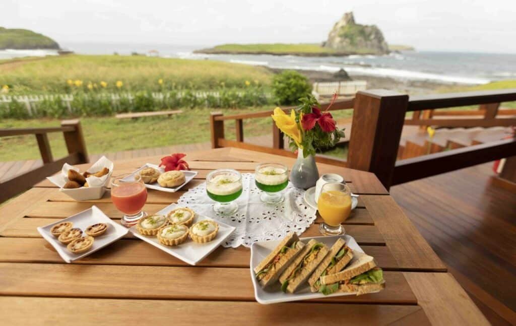 Café da manhã com lanches integrais, tortinhas de limão, outro tipo de tortinhas e mais opções em tigelas separadas, gelatina verde e suco rosa e suco amarelo, posto em mesa com vista para o mar e o jardim