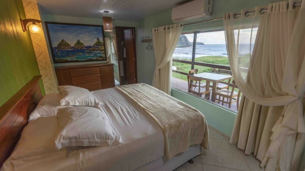 Quarto de casal na Pousada Naonda, uma das principais pousadas em Fernando de Noronha, com cama de casal, janela enorme em frente à cama, cortina blecaute e voil, vista para o mar, quadro decorativo e ar-condicionado