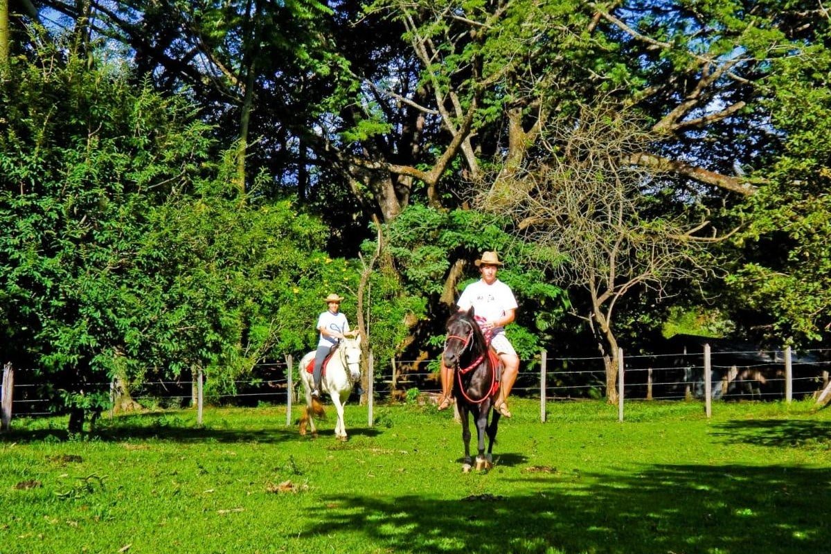 Duas pessoas cavalgam lado a lado em uma área gramada cercada por árvores na Pousada Nascentes da Fortaleza. A pessoa à esquerda cavalga um cavalo branco; a outra, um cavalo preto.